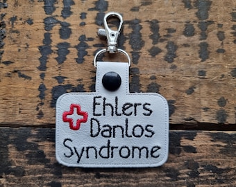 Puede incluir: Un llavero rectangular blanco con el texto "Ehlers Danlos Syndrome" bordado en negro. Una cruz roja está bordada en el lado izquierdo. El llavero tiene un cierre plateado y un botón negro.