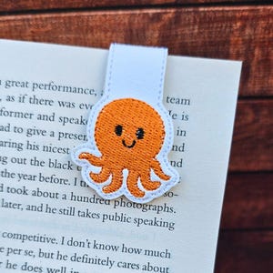 May include: An orange octopus bookmark with a smiling face, embroidered on white fabric. The bookmark is placed on the pages of a book. The octopus has eight tentacles and is a cute, whimsical design.