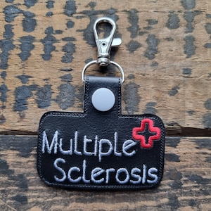 Może przedstawiać: Czarny brelok z wyhaftowanymi białymi słowami "Multiple Sclerosis". Po prawej stronie znajduje się czerwony symbol krzyża. Brelok ma srebrny zatrzask i szary guzik.