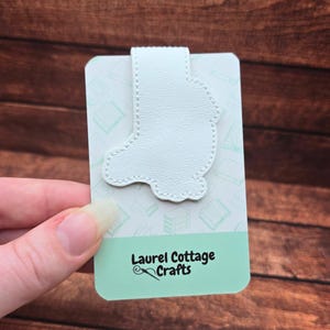 May include: A white leather bookmark in the shape of a boot is attached to a light green rectangular card with a book pattern. The card has the text "Laurel Cottage Crafts" in black. The bookmark is held by a hand.