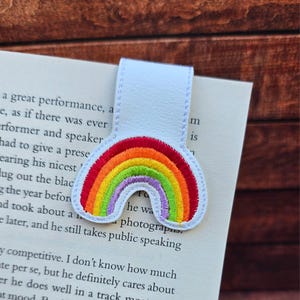 May include: A white leather bookmark featuring a stitched rainbow design in red, orange, yellow, green, and purple. The bookmark is placed in a book with printed text.