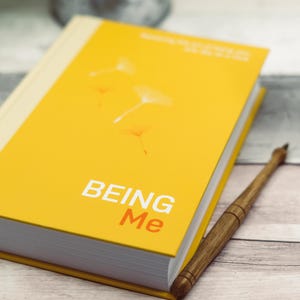 Può includere: Un libro rilegato in giallo con il titolo "BEING Me" in lettere bianche e arancioni. La copertina del libro presenta un'immagine stilizzata di semi di tarassaco che volano nel vento.