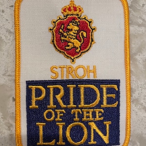 May include: A white embroidered patch with a gold border. The patch features a red and gold lion crest with a crown above it. The text "STROH" is below the crest. The text "PRIDE OF THE LION" is below the text "STROH" in a blue and gold rectangle.