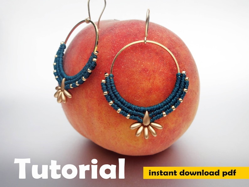 May include: A pair of gold hoop earrings with a teal blue macrame design and a gold starburst charm. The earrings are displayed on a peach.