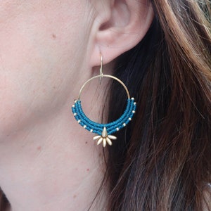 May include: Teal and gold hoop earrings with a flower design. The earrings are made of woven thread and have a gold-toned metal hoop.