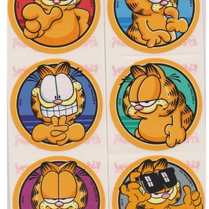 May include: Six round stickers featuring Garfield the cat in different poses and expressions. Each sticker has a colorful background and the text "© 2023 Paws, Inc." and "#1797".