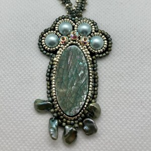 May include: A silver and green beaded necklace with a pendant featuring a large oval-shaped iridescent stone surrounded by smaller pearls and beads.