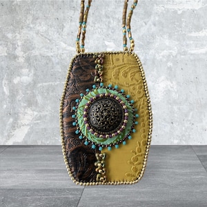 May include: A handcrafted pendant necklace with a unique design. The pendant features a combination of brown and yellow textured leather, adorned with a central circular embellishment in green, purple, and bronze. The necklace has a beaded chain.