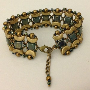 以下が含まれることがあります： 幾何学模様のゴールドトーンブレスレット。正方形のティール色のビーズと、ゴールドカラーの半月型アクセントが特徴です。小さな濃い緑色のビーズがデザイン全体に散りばめられています。ブレスレットは留め金とチェーンエクステンダーが付いています。