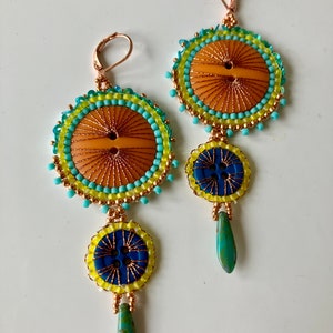 May include: A pair of dangle earrings with a blue, yellow, and orange beaded design. The earrings feature a large round button with a smaller button dangling below it. The earrings are finished with a turquoise teardrop.