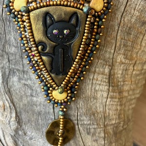 May include: A black cat pendant necklace with gold and iridescent blue beads. The cat is in a triangular frame with gold accents. The necklace has a gold button and a tassel.