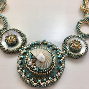 May include: An ornate necklace featuring a nautilus shell pendant and circular beaded accents in teal, gold, and white. The necklace has a detailed, handcrafted appearance, with small beads and pearl embellishments.