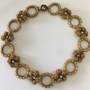 May include: A gold and white beaded necklace with a floral design. The necklace is made of small, round beads that are strung together to create a delicate and intricate pattern. The necklace is finished with a clasp that is made of metal.