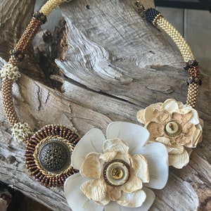 May include: A beaded necklace with a brown, gold, and white color scheme. The necklace features two large white fabric flowers with brown buttons in the center. The necklace is on a wooden surface.