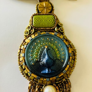 May include: A gold and green beaded necklace with a peacock pendant. The pendant features a blue and green glass cameo with a peacock design. The necklace has a gold and white pearl accent.