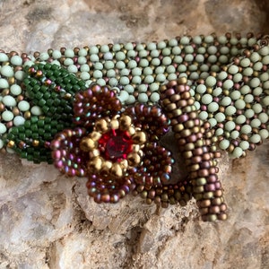 May include: A beaded bracelet with a flower design. The flower is made of brown, red, and gold beads and has a red rhinestone in the center. The bracelet is made of green and brown beads.