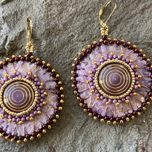 May include: A pair of purple and gold earrings with a circular design. The earrings are made with beads and fabric and have a layered, textured look.