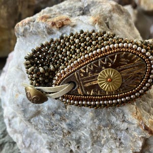 May include: A beaded bracelet with a brown leather strap and a decorative metal charm. The charm has a gold finish and a sunburst design. The bracelet is made with green, brown, and white beads.