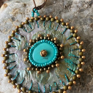 May include: Close-up of a handmade earring with a turquoise center and intricate beadwork. The earring features a circular design with a mix of green, gold, and turquoise beads, and a bronze-colored clasp. The earring is set against a textured, light-colored background.