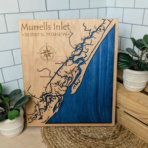 May include: A wooden map of Murrells Inlet, South Carolina, with the coordinates 33.5510° N, 79.0414° W. The map is painted blue and brown, with a compass rose in the upper left corner.