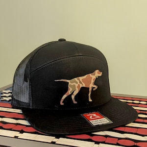 May include: Black and camo patterned flat bill trucker hat with a camo patterned dog silhouette on the front. The hat has a black mesh back and a Richardson logo on the side. "Flat Bill Snapback" is printed on the hat.