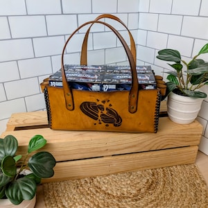 May include: A handcrafted, tan leather tote bag with two handles, filled with boxes of 12-gauge shotgun shells. The bag features a decorative burned-in design and black stitching along the sides. The bag is sitting on a wooden crate.