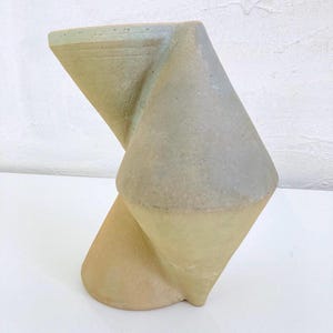 Op de afbeelding: Een abstracte keramische sculptuur met een gedraaid, geometrisch ontwerp. Het stuk heeft een mix van gedempte kleuren, waaronder beige, grijs en een vleugje groen. De vorm bestaat uit elkaar kruisende driehoekige vormen, waardoor een visueel intrigerend kunstwerk ontstaat.