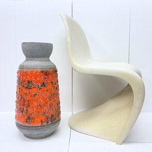 Op de afbeelding: Een keramische vaas met een getextureerd oranje en zwart ontwerp, een grijze bovenkant en een witte, gebogen stoel. De vaas heeft een uniek, ruw oppervlak. De stoel heeft een modern, minimalistisch ontwerp.