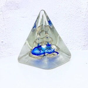 Può includere: Un fermacarte a forma di piramide in vetro trasparente con un design interno blu e argento. La forma a piramide ha una parte superiore appuntita e una base larga. Il design blu è concentrato alla base, con accenti argentati all'interno.