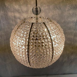 Op de afbeelding: Een bolvormige kroonluchter bestaande uit talrijke heldere, facetgeslepen kristallen kralen. De kralen zijn aan elkaar geregen om een bolvorm te vormen, met donkere metalen accenten. De lamp hangt aan een zwart snoer en wordt van binnenuit verlicht.