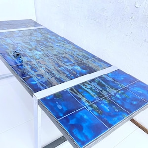 Könnte beinhalten: Rechteckiger Tisch mit blauer Fliesenoberfläche und silbernem Metallrahmen. Die Fliesen haben ein blaues und graues abstraktes Design. Der Tisch ist ca. 122 cm lang und 41 cm breit.