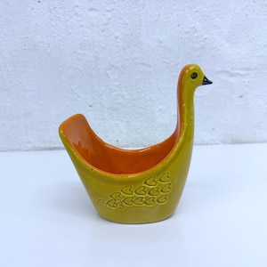 Könnte beinhalten: Eine vogelgeformte Schale aus Keramik mit einer glänzenden senfgelben Außenseite und einer leuchtend orangefarbenen Innenseite. Der Vogel hat einen langen Hals, einen kleinen schwarzen Schnabel und ein geprägtes Herzmuster an der Seite.
