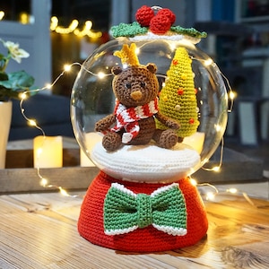 Puede incluir: Un globo de nieve navideño de crochet con un oso marrón que lleva una bufanda roja y blanca, un árbol de Navidad verde y un lazo verde en la base.