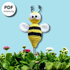 May include: A handmade crochet bee with yellow and black stripes, white wings, and black antennae. The bee is on a wooden stick, set against a backdrop of green foliage and flowers. The image includes a white circle with the text "PDF Anleitung".