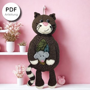 Könnte beinhalten: Eine handgefertigte braune Häkelkatze mit weißem Gesicht, rosa Nase und schwarzen Schnurrhaaren. Die Katze hat eine Tasche, in der sich eine graue Häkelmaus befindet. Der Schwanz der Katze ist gestreift, und der Text "PDF Anleitung" befindet sich in einem weißen Kreis.