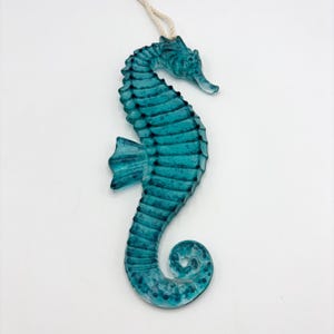 May include: A teal seahorse ornament crafted from translucent glass with a textured, segmented body and a curled tail. The seahorse features a small fin on its side and is suspended by a thin, light-coloured rope attached to the top of its head.