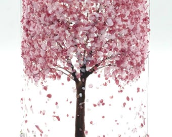 May include: A cylindrical clear glass vase featuring a fused glass design of a tree with a dark brown trunk and branches. The canopy is composed of numerous small, textured pink and white glass fragments, with scattered petals at the base.