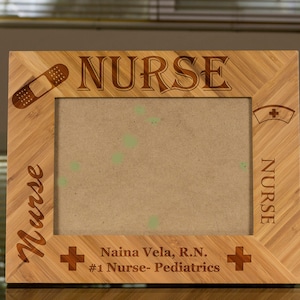 Może przedstawiać: Drewniana ramka na zdjęcia z wygrawerowanym słowem "NURSE". Ramka ma miejsce na zdjęcie i mały obrazek plastra. Tekst "Nurse" jest wygrawerowany pionowo po lewej stronie ramki. Tekst "Naina Vela, R.N. #1 Nurse- Pediatrics" jest wygrawerowany na dole ramki.