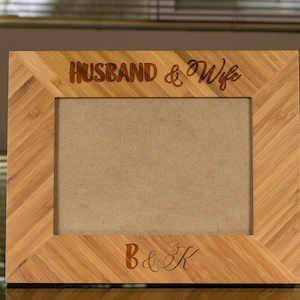 May include: A wooden picture frame with a natural bamboo finish. The frame has a beveled edge and a cutout for a photo. The text "Husband & Wife" is engraved on the top of the frame and the text "B & K" is engraved on the bottom of the frame.