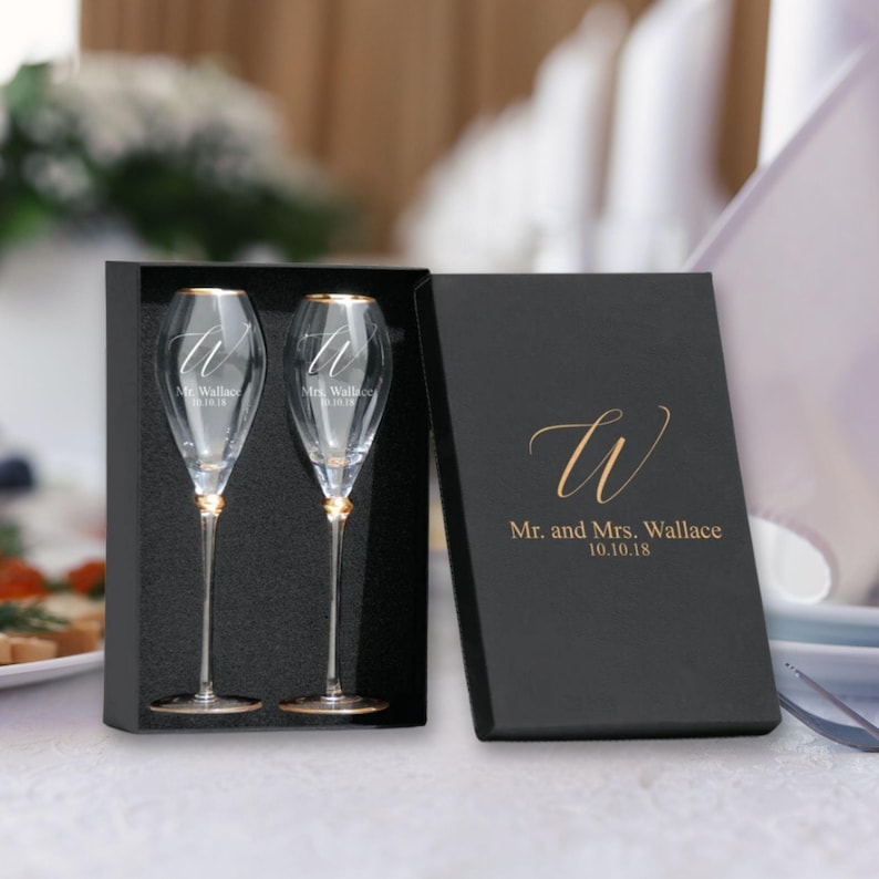 May include: Two champagne flutes with gold rims in a black box. The flutes are engraved with the letter "W" and the text "Mr. and Mrs. Wallace 10.10.18".