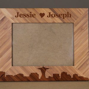 May include: A wooden photo frame with a natural wood finish and a diagonal grain pattern. The frame has a cutout of the Seattle skyline with the Space Needle at the center. The top of the frame is engraved with the text "Jessie ♥ Joseph".
