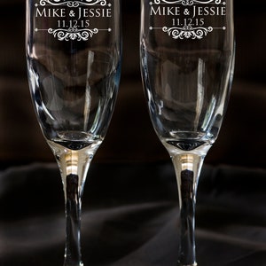 May include: Two clear glass champagne flutes with a decorative design and the text "Mike & Jessie 11.12.15" etched on the front.