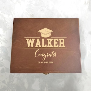 May include: A brown wooden box with a graduation cap design and the name "WALKER" in bold letters. The word "Congrats!" is written in cursive below, with "CLASS OF 2026" at the bottom. The box has a rectangular shape and a metal clasp.