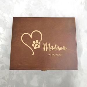 Könnte beinhalten: Eine Holzkiste mit einem Herz- und Pfotenabdruck-Design. Der Name "Madison" und die Jahreszahlen "2005-2022" sind auf dem Deckel eingraviert. Die Kiste ist mittelbraun.