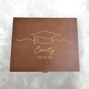 Puede incluir: Una caja de madera marrón medio con un diseño de birrete y el nombre "Christy" y "CLASS OF 2026" grabados en la tapa. La caja tiene un cierre sencillo.