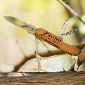 May include: A wooden handled multi-tool with a silver blade and a variety of tools. The handle is engraved with the text "Father of the Groom 6.26.17".