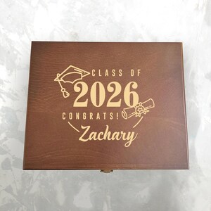 May include: A brown wooden box with a hinged lid. The box is engraved with the words "CLASS OF 2026 CONGRATS! Zachary" along with a graduation cap and diploma design in a gold color.