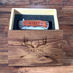 May include: A wooden box with a personalized engraved design featuring a deer's antlers and arrows. The box contains a pocket knife with a wooden handle engraved with the name "AARON".