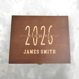 May include: A wooden box with a dark brown finish. The text "2026 class of" and the name "JAMES SMITH" are displayed in a light brown color on the box's surface. The box has a small metal clasp.