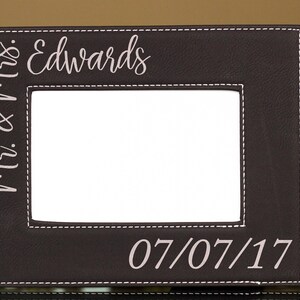 May include: A brown leather picture frame with white stitching. The frame has a white background with the text "Mr. & Mrs. Edwards" and "07/07/17" in a light gray color.